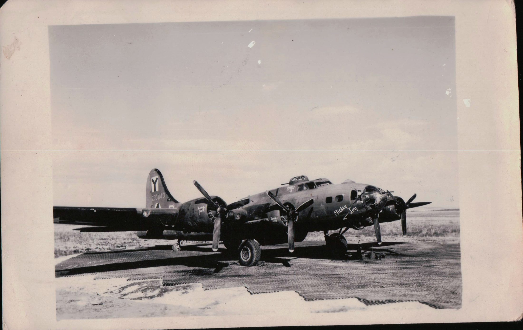 Olive drab B-17F parked on pierced steel matting, three-quarter view from the front left. The name Mickey Joy is painted below the cockpit, the tail carries a Y marking above the serial 230443, and small nose art appears near the rear fuselage.